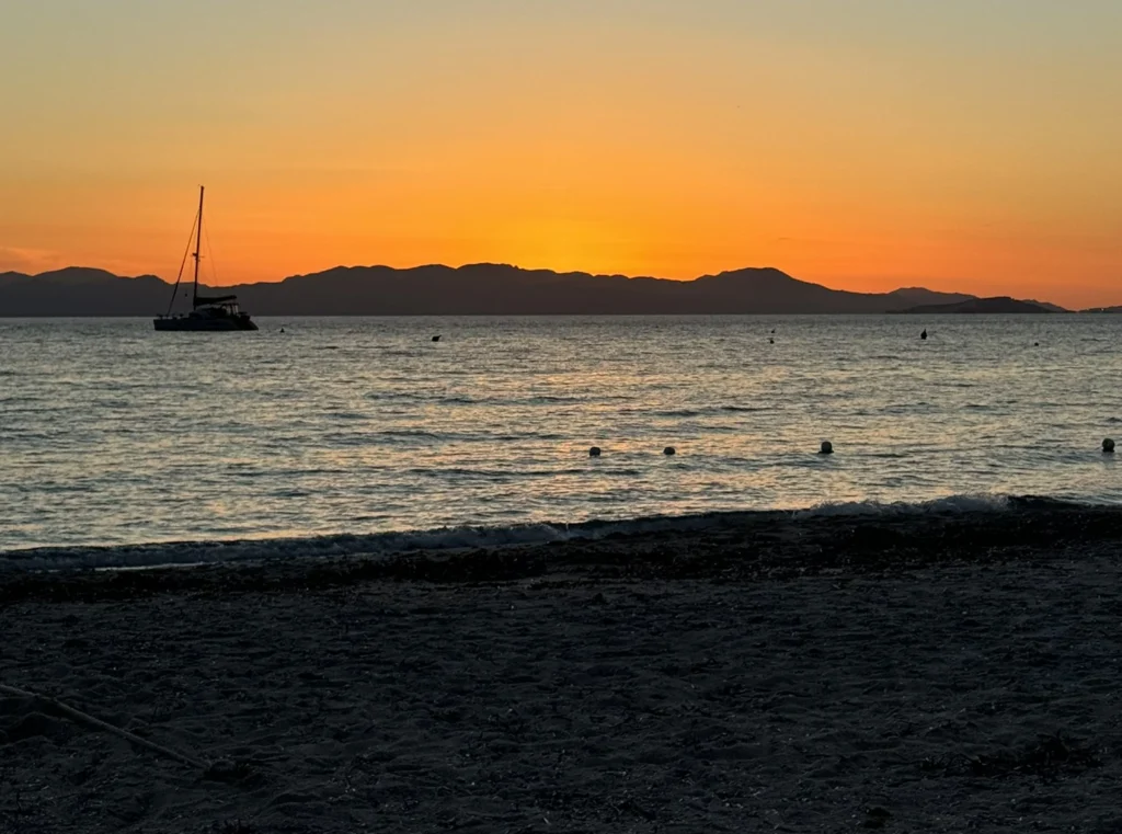 Sunset over ocean with sailboat and distant mountains.